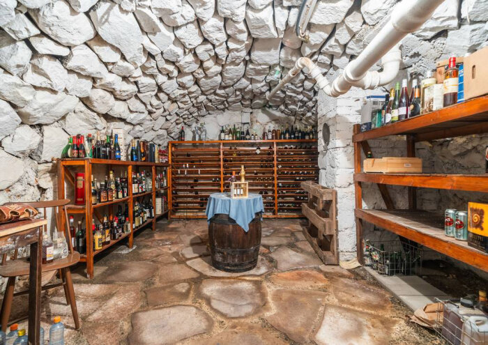 Stenkällare med välvt tak, hyllor fyllda med flaskor och en tunna med duk i mitten. Rör löper längs taket.