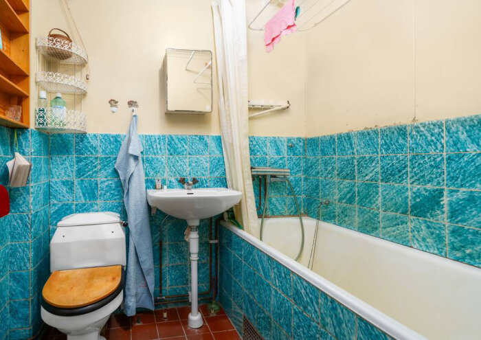 Badrum med blå kakelplattor, vit porslinstoalett med träsits, handfat med spegelskåp ovanför och ett badkar med duschdraperi.