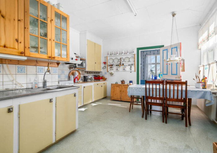 Kök med vita och träköksluckor, diskbänk, kakel med blå motiv, matbord med fyra stolar klädda i tyg och en hylla med porslin.