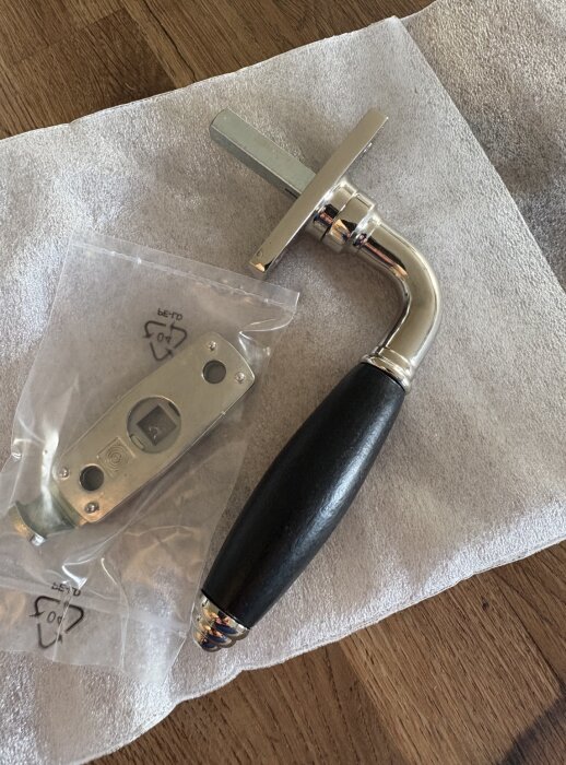 Fönsterhandtag med svart grepp och kromdetaljer bredvid en låskista i plastpåse på ett bord.