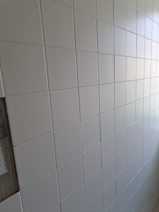 Vit kakelvägg med jämna kvadratiska plattor, en del av väggen täckt av puts eller ofärdigt område i vänsterkant.