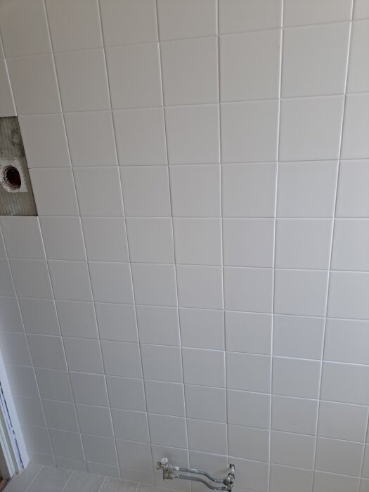 Vit kaklad vägg, jämn yta med vattenpass, vattenrör synligt i botten, trasigt kakel ovanför visar konstruktionen bakom.