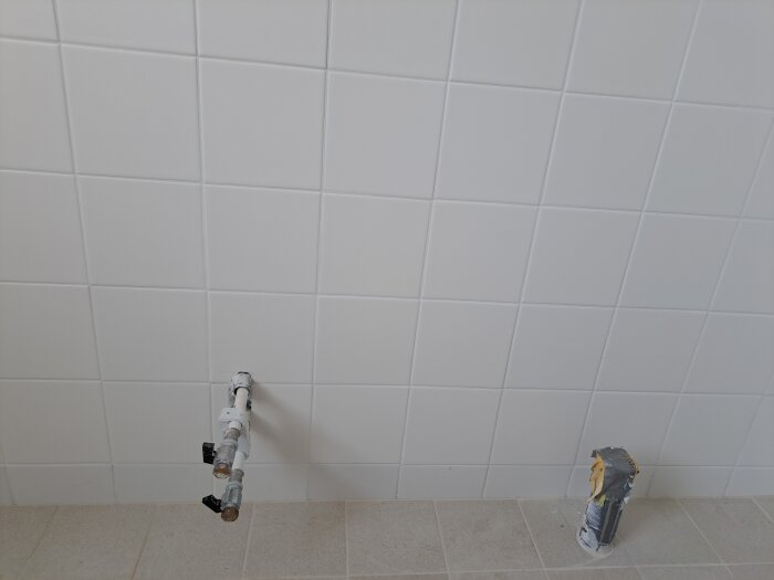 Vit kaklad vägg med exponerade rör och golvbrunn.