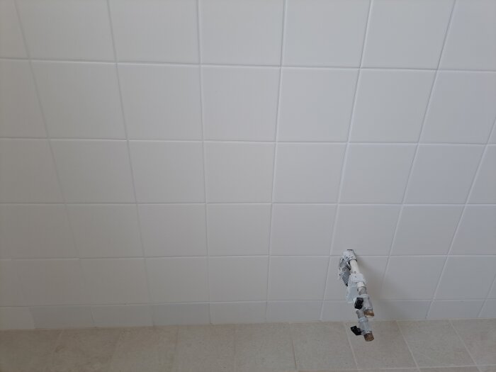 Vit kakelvägg med rör utstickande, jämn yta som fungerar bra med vattenpass, golv i ljusare kakelton.