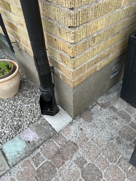 Tegelväggens hörn utifrån med en svart stupränna och stensatt mark, omgiven av en kruka med växt.