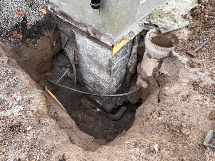 Grävt hörn av en byggnad med synlig grund, rör och jord. Del av renoveringsprojekt där fuktproblem upptäckts.
