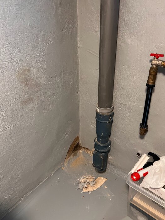Fuktfläck och skadad vägg vid rör i hörn, med rester av upplöst material på golvet.