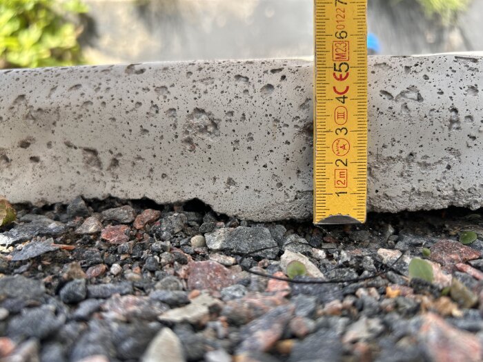 Betongplatta på grusunderlag med en gul måttstock som visar plattans tjocklek, mätt till cirka 50 mm.