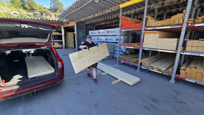 Person lastar träbrädor i en bil vid ett byggmateriallager. Förvaringshyllor med plankor och virke syns i bakgrunden.