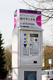 Parkeringsautomat för mobilbetalning på en trottoar med träd och bilar i bakgrunden.