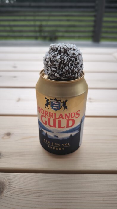 Ölburk av märket Norrlands Guld med en chokladboll täckt av kokos på toppen, placerad på ett träbord utomhus.