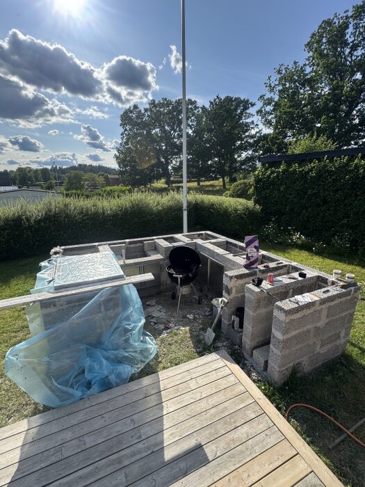 Murade block i trädgård för grillplats med gjuten skiva under arbete. Solig dag med träd i bakgrunden och verktyg synliga.