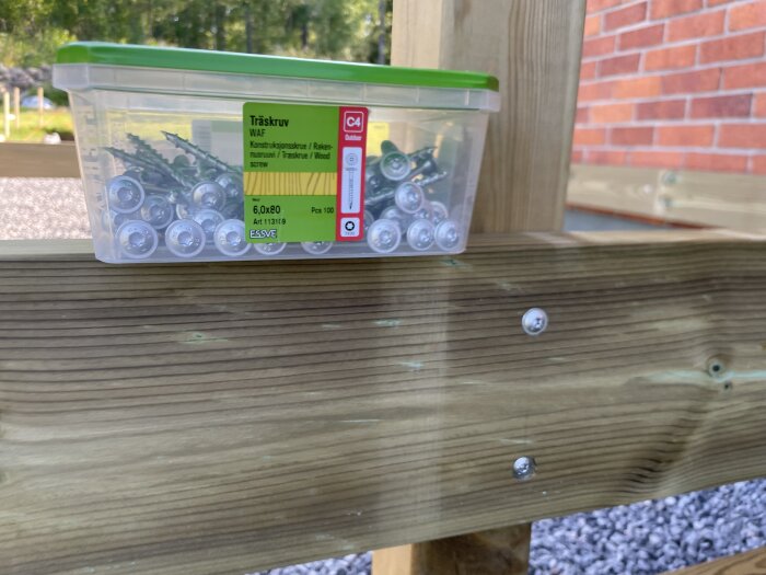 Plastlåda med träskruv WAF 6x80 på träkonstruktion utomhus, bredvid två skruvar i träet.