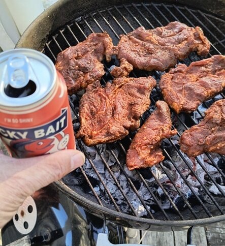 Grill med kryddade köttbitar och en öppnad ölburk i handen.