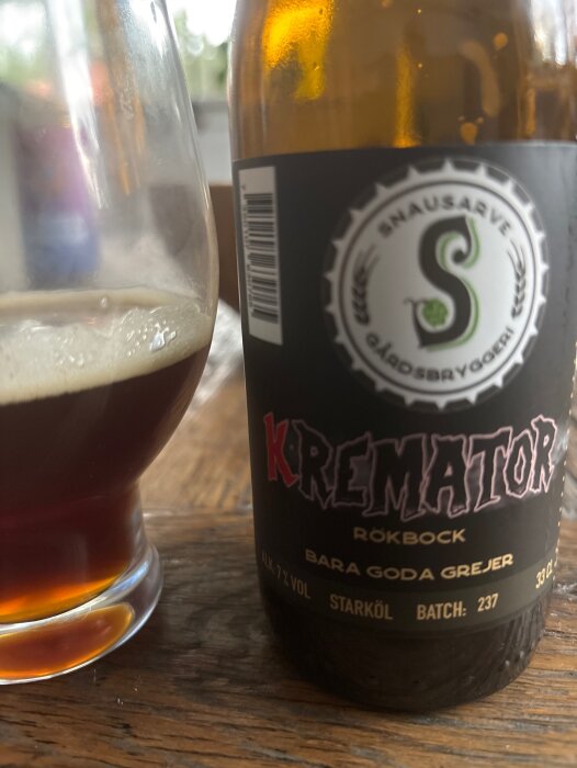 Flaska och glas med rökölen "Kremator Rökbock" från Snausarve Gårdsbryggeri på ett träbord.