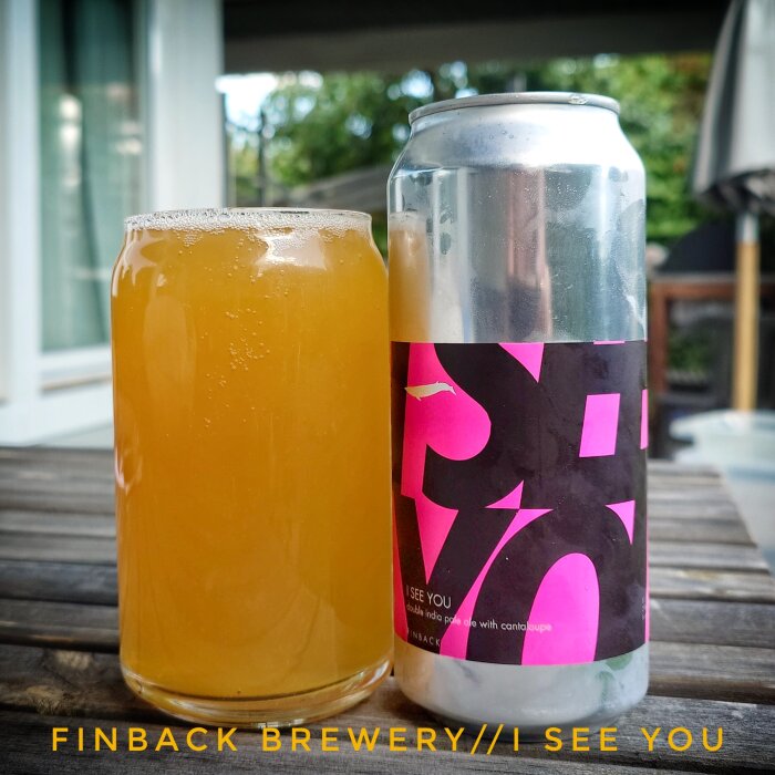 Ölglas med gul dryck bredvid burk från Finback Brewery med etikett "I See You", stående på ett bord utomhus.