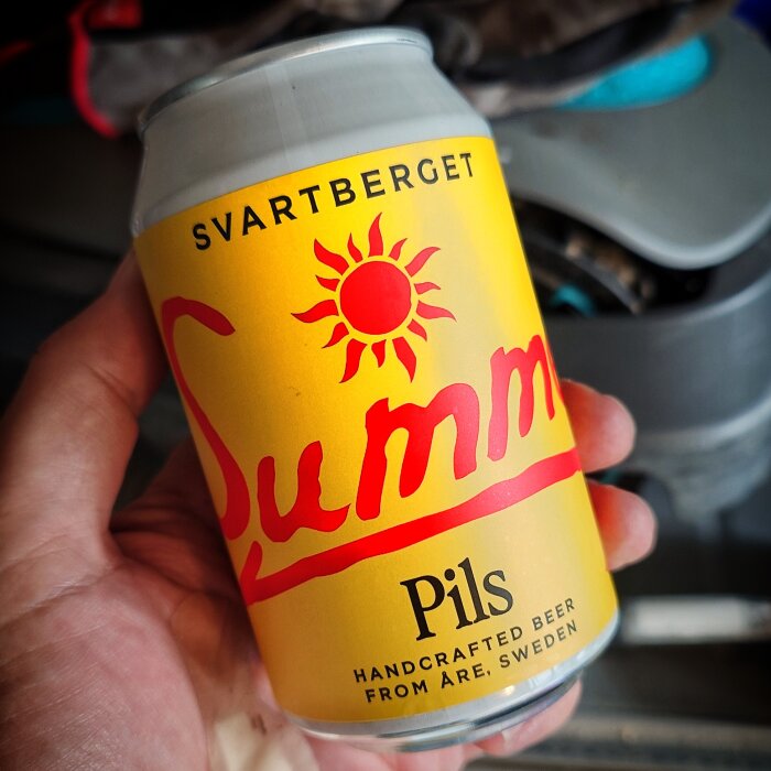 Hand som håller en gul ölburk med texten Svartberget Summer Pils och en solsymbol, från Åre, Sverige.