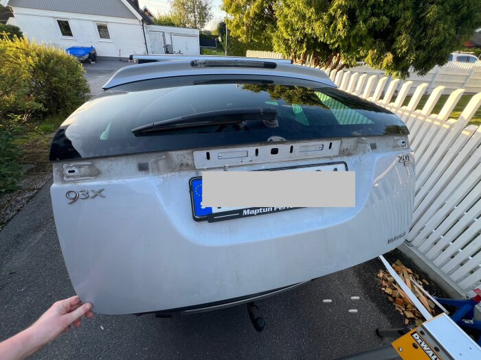 Bilens baklucka och nummerskylt utan lampa visas, omgiven av verktyg på marken. Nya glödlampkontakter tillverkas av metallband.