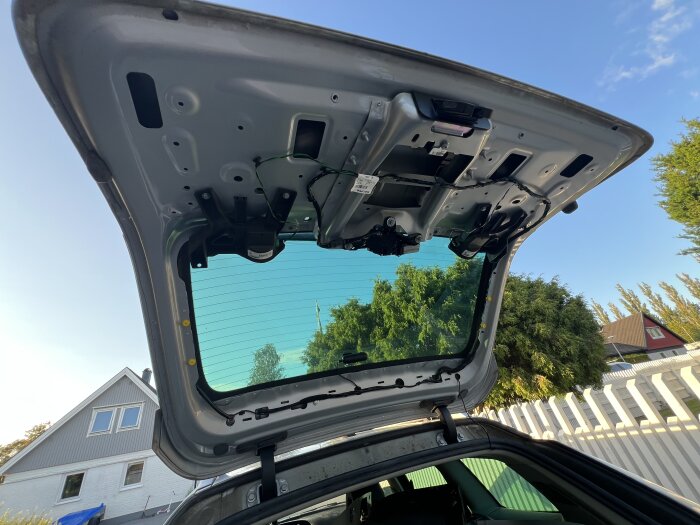 Öppen baklucka på en bil, visar nummerskyltens belysning och ledningar, omgiven av hus och träd i bakgrunden.