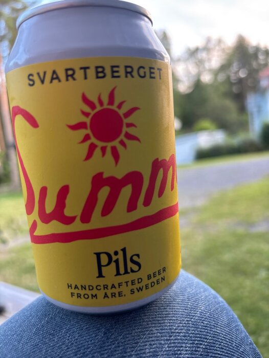 En gul och röd burk med "Svartberget Summer Pils" öl från Åre, Sverige, uppställd utomhus med en grönskande bakgrund.
