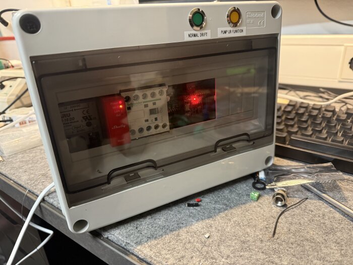 Självbyggd styrbox för pumpautomation med Shelly-modul och relä, monterad på en arbetsbänk. Två knappar märkta "Normal drift" och "Pump ur funktion".