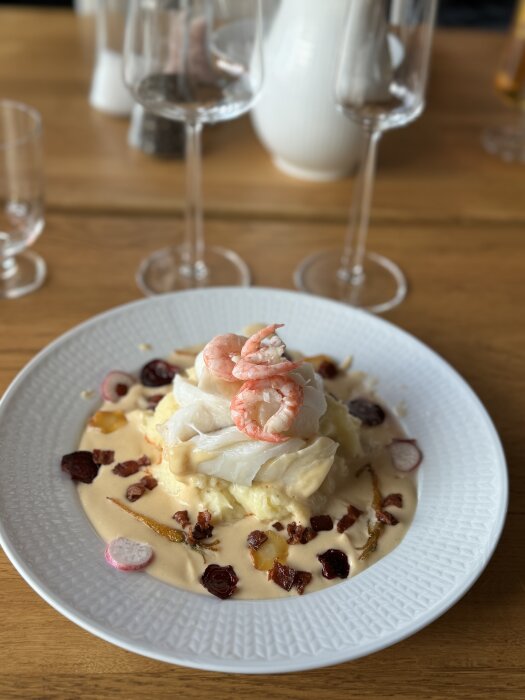 Tallrik med pocherad torskrygg och räkor i en krämig sås, serverad med grönsaker och krispiga bitar, på en träbord.