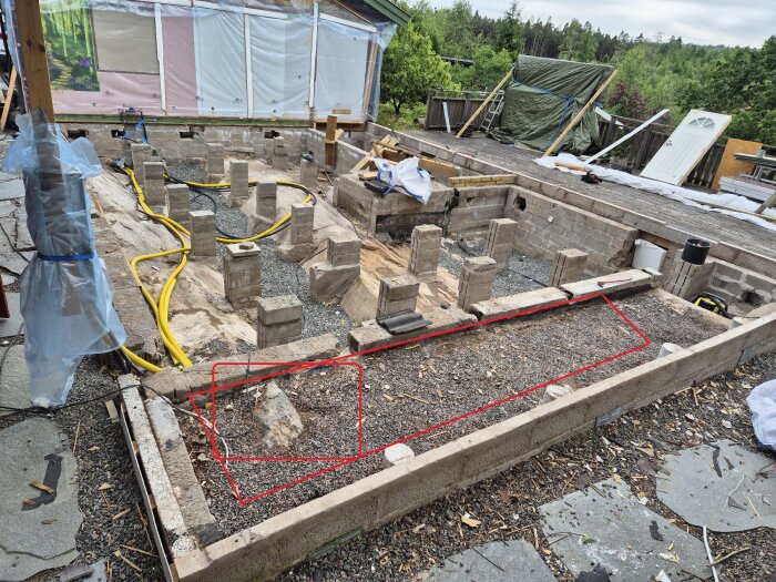 Grunden med plintar av betong och grus, med röda markeringar över ovälkommet berg som sticker upp. Grönska och byggmaterial i bakgrunden.