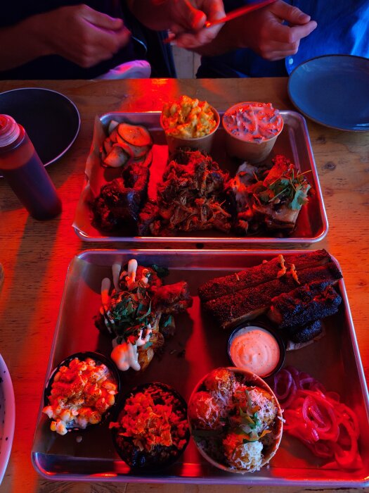 Tre serveringsbrickor med grillat kött, tillbehör som mac n' cheese och potatis, samt tre såsskålar. Två personer äter vid ett träbord.