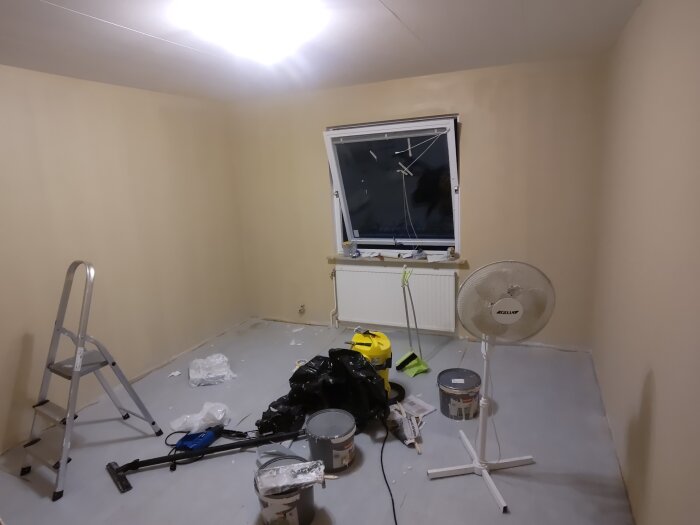 Rum under målning med beige färg, stege och fläkt. Målarburkar och verktyg på golvet, öppet fönster i bakgrunden.
