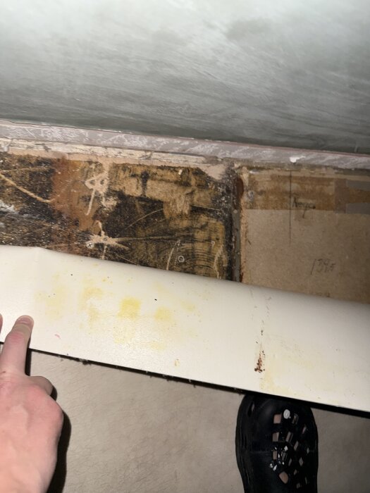 Plastmatta lyfts upp i ett hörn för att visa gammalt svart lim på golvet under, en hand pekar på mattkanten.