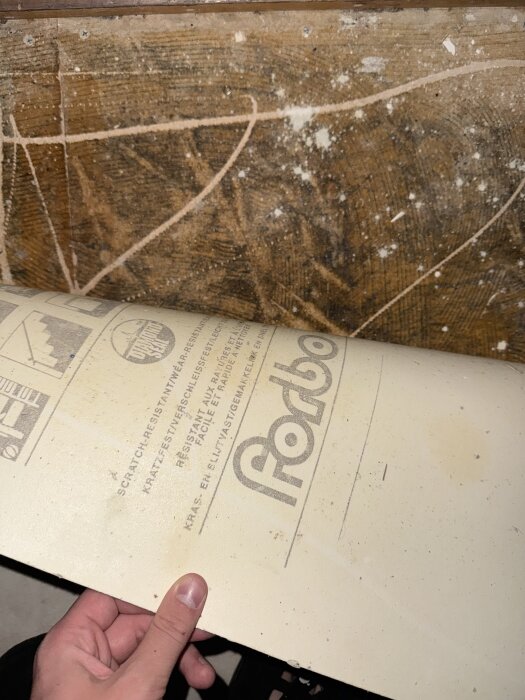 Plastmatta lyfts upp från golv för att visa gammalt svart lim under, med texten "Forbo" synlig på baksidan av mattan, misstänkt för analys.