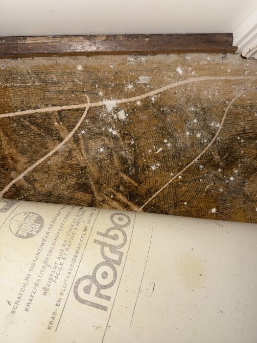 Plastmatta från Forbo lyfts upp, avslöjande gammalt svart lim och slitage på trägolvet under.