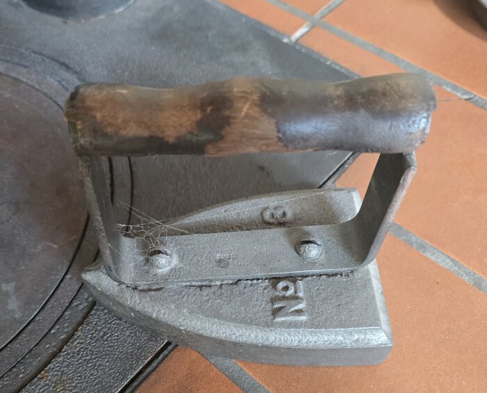 Gammaldags strykjärn med trähandtag på ett köksfat av gjutjärn och terracotta golvplattor.