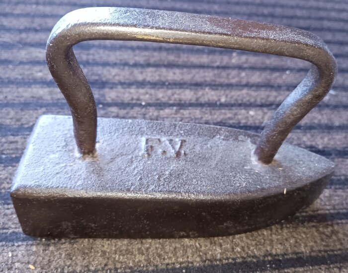 Järnföremål från 1700-talet med handtag, märkt med "FV", oklart ursprung, placerat på randigt underlag.