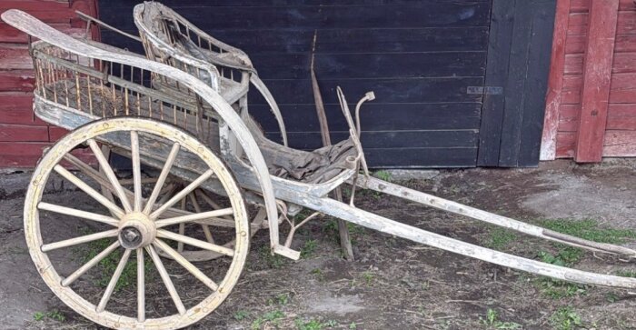 En gammal trälastkärra med två stora hjul, placerad utanför en ladugårdsdörr på en gårdsplan.
