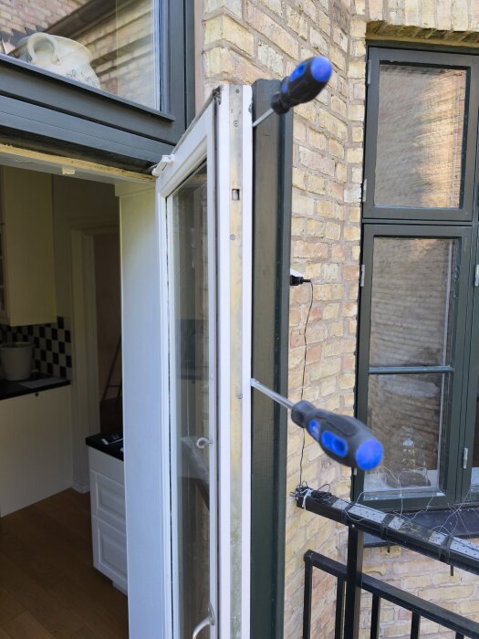 Balkongdörr i äldre hus med två skruvmejslar som försöker öppna den, visar tegelvägg och insidan av bostad genom öppet hål.