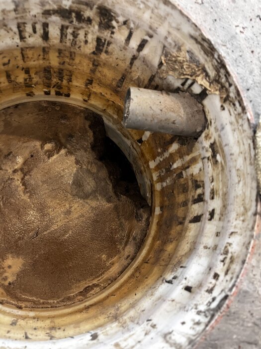 Rund pumpbrunn med synlig betongbotten och ett rör som går in i brunnen, omgiven av missfärgad betong och sediment.