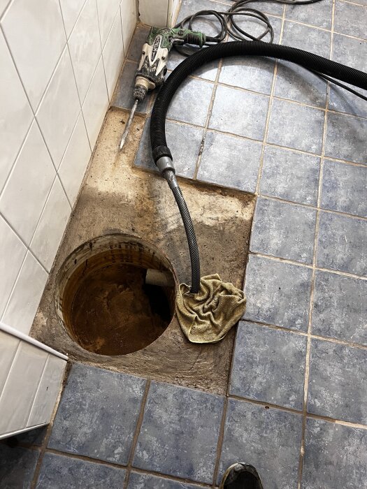 Öppen brunn i klinkergolv med borr, vakuumslang och trasor runt, del av ett källarprojekt för att sänka vattennivån och gjuta botten.