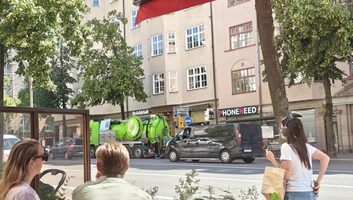 Tre personer sitter utomhus vid ett café. En grön slamtömningsbil syns på gatan framför en beige byggnad.