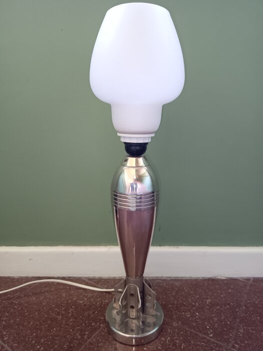 Äldre bordslampa med metallfot, liknar en projektil, med vit lampskärm och grön bakgrund. Eventuellt gjuten i Eskilstuna, Luth&Rosen.