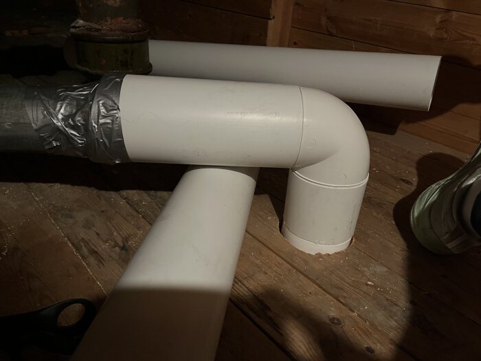 Plaströr och metallrör i ett badrum, anslutna med tejp, del av ventilationssystem för paxfläkt. Trägolv och metallbeslag syns.