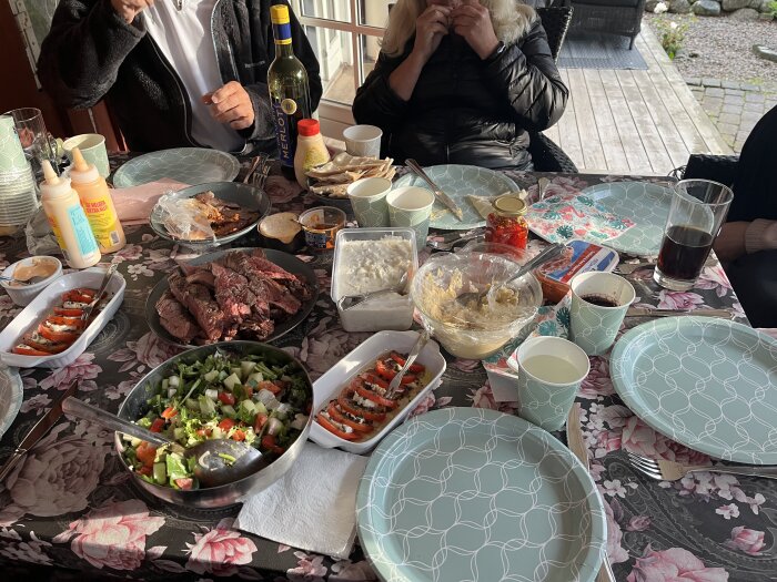 Matbord med olika rätter: kött, sallad, såser och drycker. Tallrikar och glas uppställda, människor runt bordet.