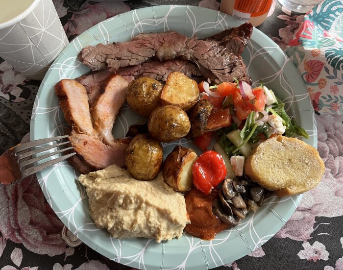 Tallrik med pluma, flapstek, potatis, sallad, bröd, hummus, svamp och röd sås på en blommig duk.