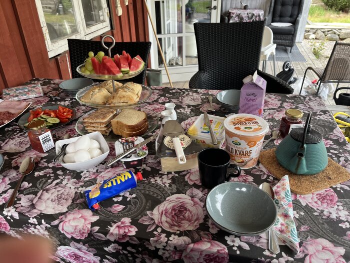 Frukostbord med bröd, fruktfat, kokta ägg, kvarg, te i kanna, brödskivor och pålägg på en blomsterduk.