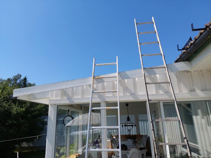 Två stegar lutade mot ett vitmålat altantak med en lätt lutning, utrustat med stora fönster och en uteplats under en blå himmel.