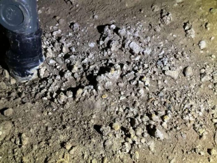 Jordig markyta i en krypgrund med inslag av grus och små stenar, delvis belyst, samt ett svart rör synligt i övre vänstra hörnet.