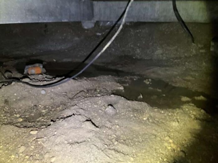 Jordgolv i krypgrund med lösa kablar och vattenpöl, del av äldre hus utan åldersbeständig plast, trossbotten synlig ovanför.