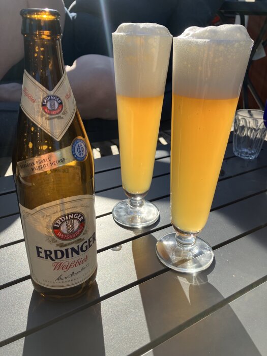 En flaska Erdinger Weißbier och två fyllda ölglas med skum på en uteplats i solljus.