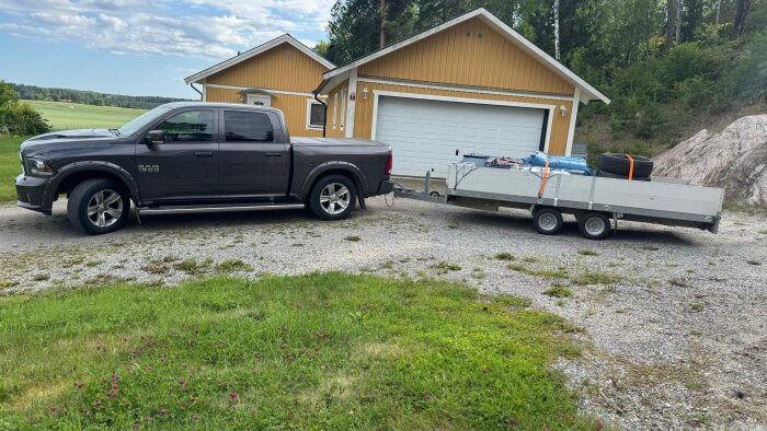 Pickup med släpvagn, lastad med däck och annat, står parkerad framför ett gult hus med dubbelgarage i lantlig miljö.