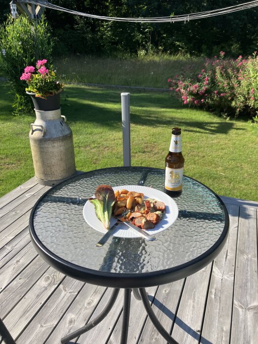 Tallrik med grillrester, potatisklyftor och sallad på glasbord utomhus, ölflaska bredvid, blommor och gräsmatta i bakgrunden.